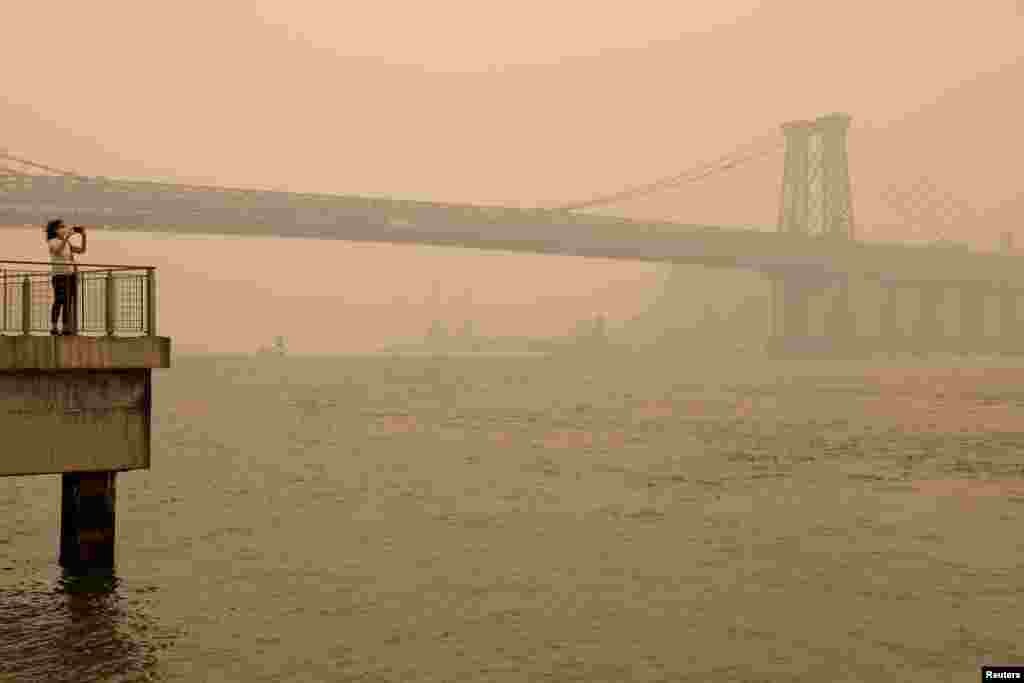 A person wearing a face mask takes a picture of the Manhattan skyline while it's covered in haze and smoke caused by wildfires in Canada, in Brooklyn, New York, June 7, 2023.