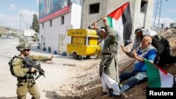 Demonstran memegang bendera Palestina di depan seorang tentara Israel yang memegang senjata saat protes menuntut Israel pembukaan kembali jalan yang ditutup menuju Nablus, di Deir Sharaf, di Tepi Barat yang diduduki Israel, 20 Oktober 2022. (Foto: Reuters)
