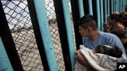 Pripadnik karavana migranata iz Centralne Amerike, držeći dete, gleda kroz pograničnu ogradu prema grupi ljudi koji su se okupili na američkoj strani, dok stoji na plaži gde se pogranična žica završava u okeanu, u Tihuani, Meksiko, 29. aprila 2018.