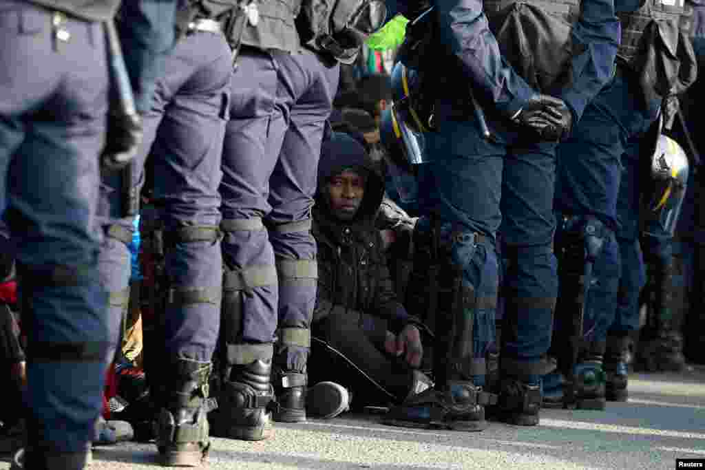 Polisi Perancis berdiri dekat para migran yang menunggu di tempat pemrosesan untuk didaftarkan pada hari kedua evakuasi dan pemindahan mereka ke pusat-pusat penerimaan di Perancis, di tengah pembongkaran kamp Calais, di Perancis (25/10). (Reuters/Neil Hall)