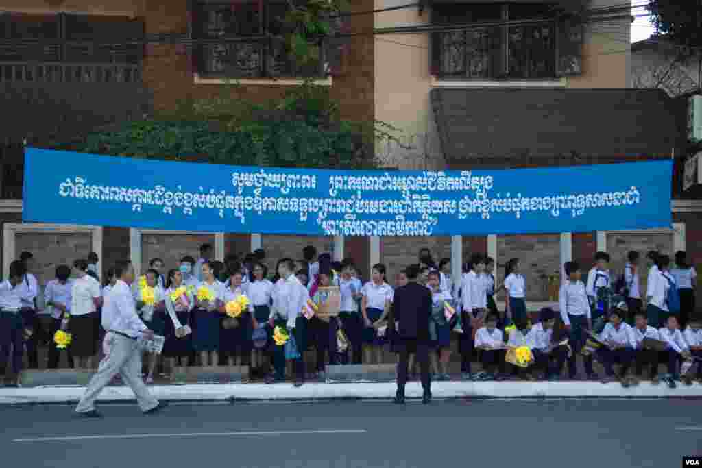 Students are waiting on the pavement under the banner with text honoring the title "Preah Sri Loka Dhammika Raja" to King Norodom Sihamoni on June 3, 2015. (Nov Povleakhena/VOA Khmer)