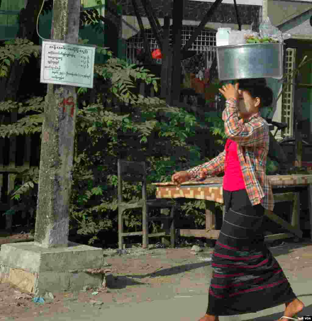 Seorang perempuan mengenakan sarung longyi dan bedak dingin thanaka di pipinya sambil membawa makanan. (VOA/Steve Herman)