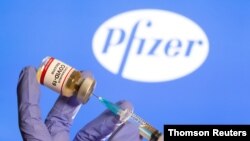 Una mujer sostiene una pequeña botella con una etiqueta que reza 'vacuna contra el coronavirus COVID-19' y una jeringa médica frente al logotipo de la farmacéutica Pfizer.