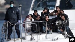 Un officier de la garde côte grecque, portent un masque contre les maladies infectieuses, se dresse à côté d'un petit groupe d'immigrants, lors d’une opération de récupération de près de 700 immigrants clandestins, au port crétois côtière de Ierapetra, Grèce, le jeudi 27 novembre 2014.
