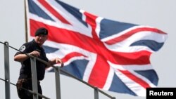 英國皇家海軍海神之子兩棲登陸艦抵達日本東京一個港口時一名英國水手站在英國國旗前。（2018年8月3日） 