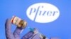 Una mujer sostiene una pequeña botella con una etiqueta que dice "Vacuna contra el coronavirus COVID-19" y una jeringuilla frente al logotipo de Pfizer. [Foto: Archivo]
