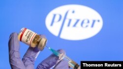 Una mujer sostiene una pequeña botella con una etiqueta que dice "Vacuna contra el coronavirus COVID-19" y una jeringuilla frente al logotipo de Pfizer. [Foto: Archivo]