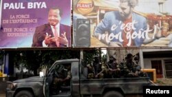 Des membres du Bataillon camerounais d'élite d'intervention rapide (BIR) assis sur leur véhicule militaire sous panneau de campagne (à gauche) du président camerounais Paul Biya, candidat à la réélection prévue le 7 octobre, lors d'une patrouille dans la ville de Buea, au sud-ouest du pays Cameroun, 4 octobre 2018.