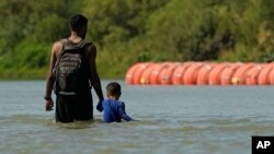 Migrantes caminan junto a grandes boyas que se utilizan como barrera fronteriza flotante en el río Bravo, el 1 de agosto de 2023, en Eagle Pass, Texas.