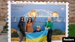 Gratë ukrainase pozojnë përpara një vepre arti në Kiev që pasqyron zjarrin në urën e Krimesë