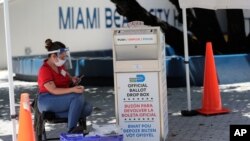Članica biračkog odbora nosi zaštitnu opremu dok posmatra glasačku kutiju sa glasovima pristiglim poštom, tokom ranog glasanja, 7. avgusta 2020, u Majami Biču, Florida.