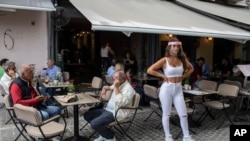 A cafe-restaurant staff stands outside as people drink coffee in Monastiraki district of Athens, on Monday, May 25, 2020. Greece restarted regular ferry services to its islands Monday, and cafes and restaurants were also back open for business as…